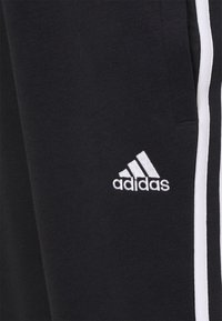 Schwarzer Stoff mit weißem, gesticktem Adidas-Logo in der Nähe von weißen vertikalen Streifen auf Sportmaterial.