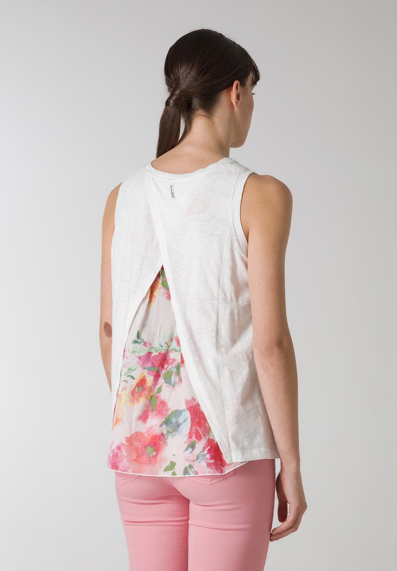 Vit ärmlös topp med blommönster på ryggen, med en delad design, bärs tillsammans med rosa byxor. Mjukt, lättviktigt material.