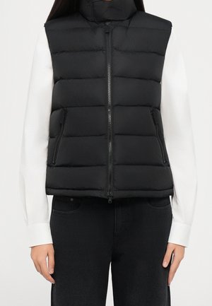 Veste noire matelassée sans manches avec fermeture éclair devant et poches zippées, portée sur une chemise blanche à manches longues et un pantalon noir.