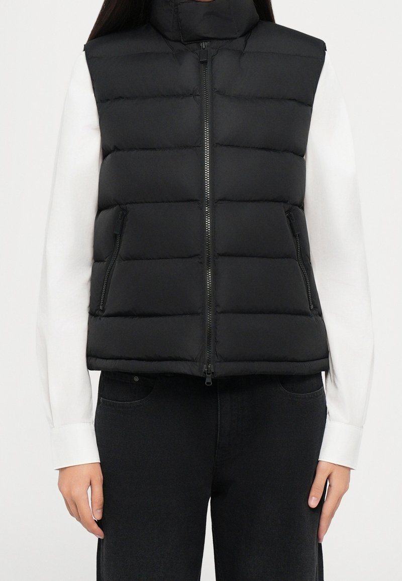 Veste noire matelassée sans manches avec fermeture éclair devant et poches zippées, portée sur une chemise blanche à manches longues et un pantalon noir.