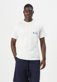 White cotton t-shirt featuring a blue and green Quiksilver logo on the left chest. Round neckline and short sleeves. Navy shorts.