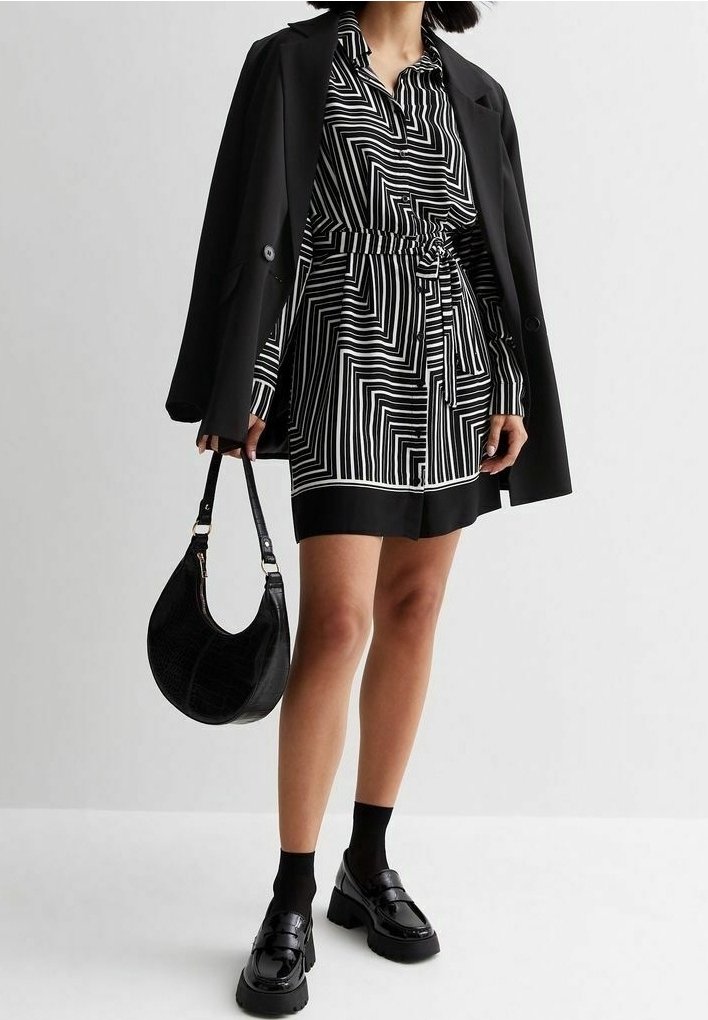 Femme portant une robe à motif géométrique noir et blanc avec un blazer noir drapé sur les épaules, tenant un sac à main noir et portant des chaussures noires.