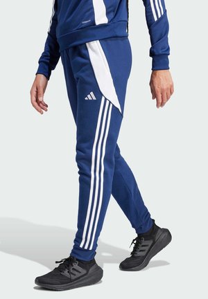Marineblaue Sport-Jogginghosen mit weißen Streifen, schmaler Schnitt und akzentuiertem Seitenpanel. Kombiniert mit schwarzen Sneakers, die eine strukturierte Sohle haben.