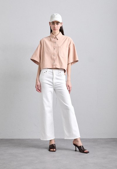 Rožinė trumpa mygtukinė palaidinė, balti plačiakraščiai džinsai, rudi atviri bateliai su kulnu ir šviesus kepuraitė, prieš neutralų foną.