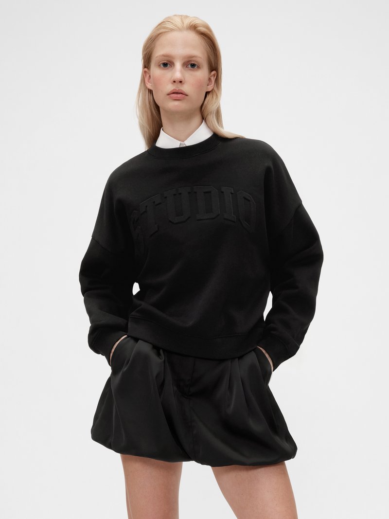 Sweatshirt noir avec texte en relief "STUDIO". Doté d'un col rond et de poignets côtelés, il est associé à un short plissé noir.