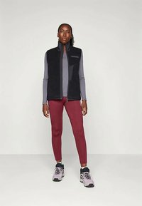 Vesta neagră din fleece cu guler înalt purtată peste o cămașă gri cu mânecă lungă, asortată cu pantaloni mulati vișinii și adidași gri.