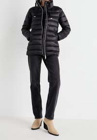 Černá prošívaná bunda s předním zipem, nastavitelnou kapucí, horizontálním prošitím a dvěma bočními kapsami. Kombinována s černými džínami a béžovými botami na podpatku.