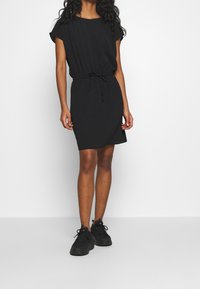 Robe noire à manches courtes avec taille à cordon, fabriquée en tissu doux, présentant une coupe décontractée et une longueur au genou. Assortie à des baskets noires.