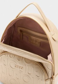 Zaino in pelle beige con logo in rilievo, chiusura a zip e interno rosa con una piccola tasca con zip. Forma arrotondata e manico superiore.