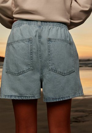 Inimene, kes kannab helesiniseid teksapükse ja beežikat ülaosa, seisab liivarannal päikeseloojangul, selg kaamerale.