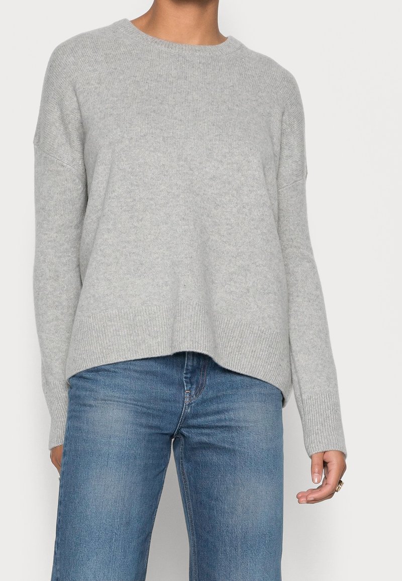 Personne portant un pull en maille gris clair à col rond et un jean bleu délavé, debout devant un fond blanc uni.