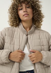 Veste bomber matelassée beige avec col côtelé et fermeture éclair sur le devant. Elle présente des manches longues et arrondies ainsi qu'une couture horizontale subtile.