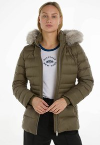 Giacca a vento verde oliva, imbottita, con cappuccio foderato in pelliccia, chiusura con zip e polsini a coste, indossata sopra una maglietta bianca con dettagli blu e logo.