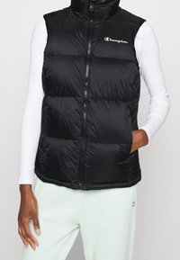 Svart pufferväst med hög krage, framdragkedja och logotyp. Tillverkad av glänsande material, med ett kviltat mönster och sidofickor.