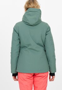 Grön huva jacka med en slät, texturerad finish och söm detaljer, parat med klara rosa byxor, sedd från baksidan.