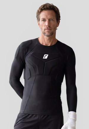 Mann trägt schwarzes gesteppte Sportshirt und weißen Handschuh vor hellgrauem Hintergrund, schaut geradeaus mit neutralem Gesichtsausdruck.