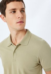 Camisa polo caqui con un cuello texturizado, que presenta una placket de botones y mangas cortas. El material parece transpirable y estructurado.