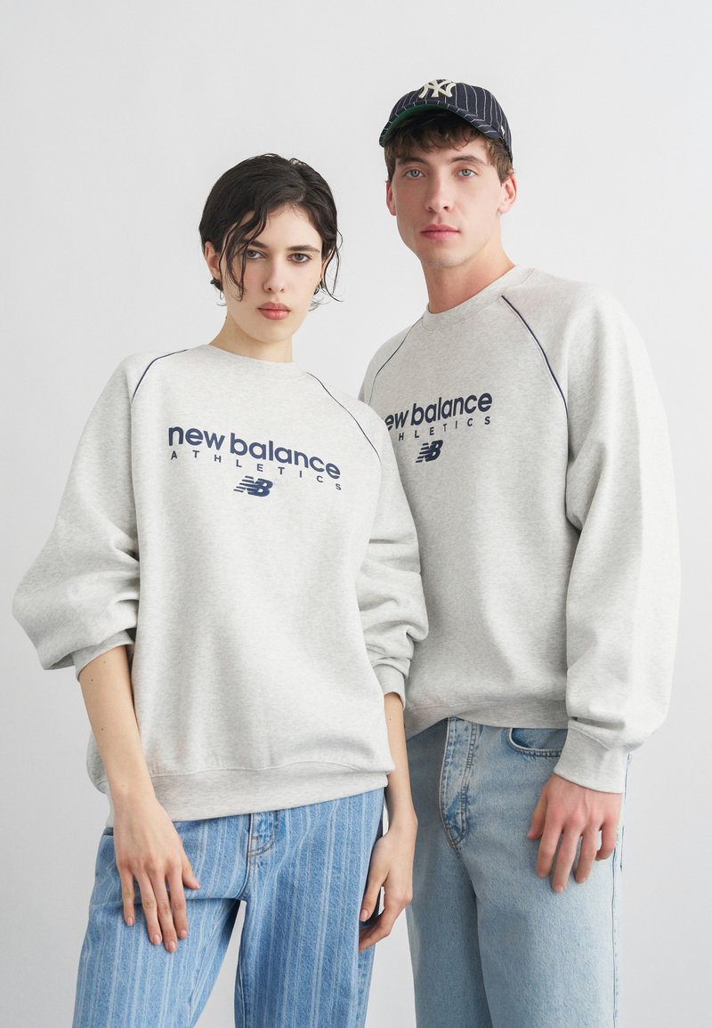 Kaksi henkilöä, jotka pukeutuvat harmaisiin New Balance Athletics -collegepaitoihin ja sinisiin farkkuihin, seisovat yksinkertaista vaaleaa taustaa vasten.