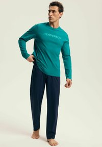 Turkusowa koszula z długim rękawem z napisem "HENDERSON", w połączeniu z ciemnogranatowymi spodniami, obie wykonane z miękkiego materiału. Model stoi boso.