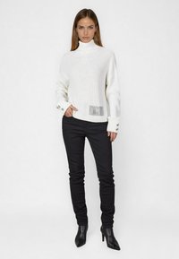 Weißer gerippter Rollkragenpullover mit einem strukturierten schwarzen Patch, verziert mit silbernen Knopfakzenten an den Ärmeln, kombiniert mit schmalen schwarzen Jeans und spitzen Stiefeln.