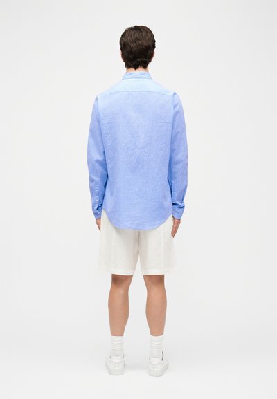 Άνδρας που στέκεται με την πλάτη, φοράει ανοιχτό μπλε μακρυμάνικο πουκάμισο, άσπρα σορτς μέχρι το γόνατο, άσπρες κάλτσες και άσπρα αθλητικά παπούτσια μπροστά σε ένα απλό φόντο.