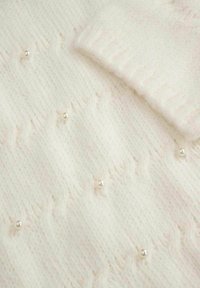 Biały dzianinowy materiał z teksturowanym wzorem i małymi perełkowymi zdobieniami. Zawiera złożony mankiet z dopasowanego dzianinowego materiału.