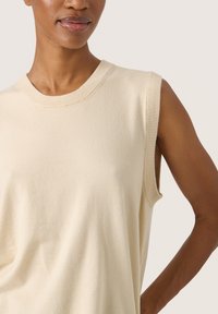 Débardeur en tricot beige sans manches avec un col rond, texture lisse, éclat subtil et fentes sur les côtés pour faciliter les mouvements.