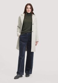 Grijze wollen jas met een enkelvoudige sluiting, groene coltrui, marineblauwe ribfluweel broek en zwarte instappers. Effen achtergrond.