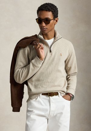 Homme portant un pull beige à zip quart, pantalon blanc, lunettes de soleil, tenant une veste marron sur l'épaule, avec montre et ceinture marron visibles.