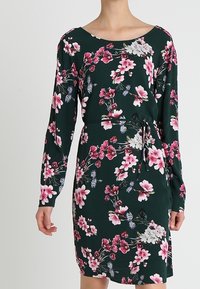 Bloemenjurk met lange mouwen, donkergroene basis, roze en witte bloemenpatroon, en een strikceintuur in de taille. Zachte stof, aansluitend ontwerp.