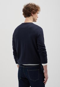 Marineblaues Strickpullover mit langen Ärmeln, gerippten Bündchen und Rundhalsausschnitt. Über einem weißen Hemd getragen, von hinten gezeigt.