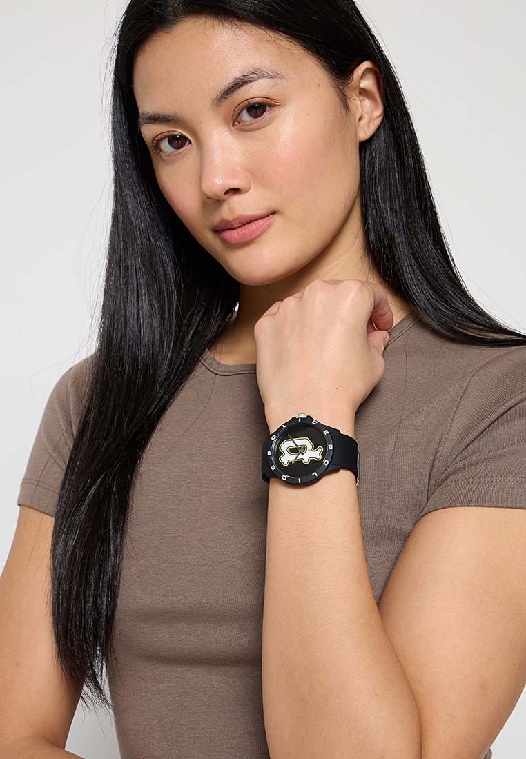 Reloj de goma negra con esfera redonda que presenta un audaz diseño gráfico en amarillo, llevado en una muñeca. Textura suave y correa ajustable.