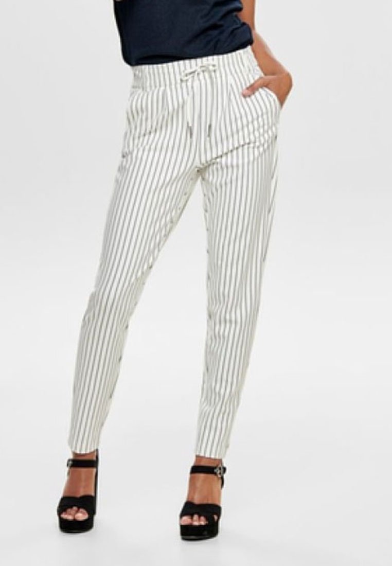 Personne portant un pantalon blanc à rayures verticales grises, des sandales noires à talons hauts, et un haut bleu marine rentré, debout avec une main dans la poche.