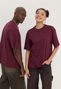 Bordo majice s kratkimi rokavi in ohlapnim krojem. Ena model nosi temne hlače, druga pa svetle kavbojke. Minimalističen dizajn, gladka tkanina.
