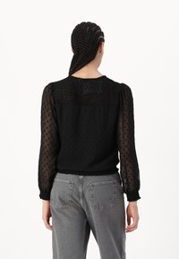 Blouse noire avec des manches transparentes à pois, un corsage texturé, une emmanchure froncée au dos et un ourlet à volants. Assortie avec un jean taille haute gris.