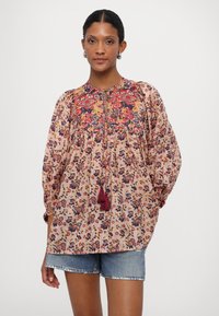 Blümchenbluse in Hellrosa mit gerafftem Ausschnitt, besticktem Schulterbereich in Rot und Marineblau sowie Quastenakzenten. Getragen mit Jeansshorts.