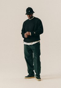 Mann trägt einen schwarzen Strickpullover, darunter ein gestreiftes Shirt, weite dunkle Hosen, einen schwarzen Hut mit Logo und lässige Schuhe, steht vor einem beigen Hintergrund.