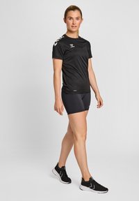 Svart trenings t-skjorte med korte ermer, med logo og kontrasterende skulderstriper, kombinert med svarte shorts og svarte sneakers.