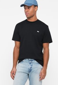 Čierne tričko s krátkym rukávom a okrúhlym výstrihom s logom Lacoste. Nosené s mierne modrými poškodenými džínsami a denimovou šiltovkou.