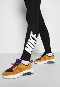 Nike sneakers w żółtym i czarnym kolorze z nakrapianą białą podeszwą, skomplikowane sznurowanie w wielu kolorach, zestawione z czarnymi legginsami z białym logo.