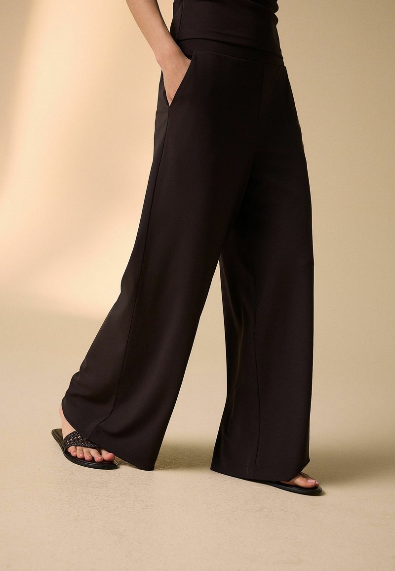 Personne portant un pantalon noir à jambes larges avec des poches, associé à des sandales noires tressées, debout sur un sol et un fond beiges.