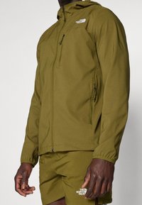 Olive green jacket with zippered front, adjustable hood, and elastic cuffs. Features a chest pocket and logo on the left side.