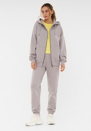 Two-piece loungewear set featuring a light grey zip-up hoodie and sweatpants, paired with a yellow t-shirt and cream sneakers.
