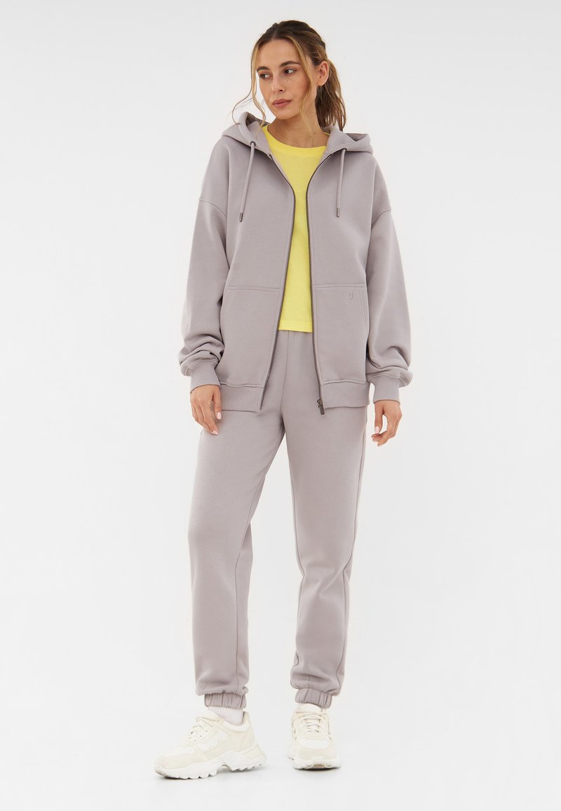 Twee-delige lounge-set bestaande uit een lichtgrijze rits-hoodie en joggingbroek, gecombineerd met een gele t-shirt en crèmekleurige sneakers.