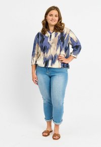 Blusa leggera e stampata in blu navy, crema e beige, con scollatura a bottoni; abbinata a jeans azzurri e sandali marroni.
