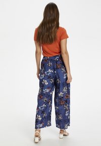 Floral patronen, wijde broeken van blauwe stof met witte, rode en lavendelkleurige bloemen. Elastische tailleband en losse pasvorm. Witte haksandalen.