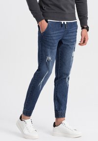 Pantalones jogger de mezclilla azul oscuro con detalles desgastados, puños elásticos y bolsillos laterales, combinados con zapatillas blancas con detalles en negro.