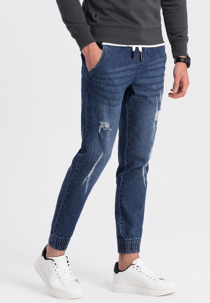 Pantalones jogger de mezclilla azul oscuro con detalles desgastados, puños elásticos y bolsillos laterales, combinados con zapatillas blancas con detalles en negro.