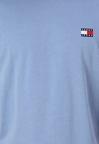 Světle modré bavlněné tričko s krátkými rukávy a malým černým, červeným a bílým logem "TOMMY JEANS" na levé straně hrudi. Hladká textura.