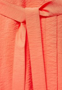 Koralu materiał teksturowany z szczegółem w postaci wiązanego paska. Tkanina ma pomarszczony wygląd, co zwiększa jej atrakcyjność wizualną.